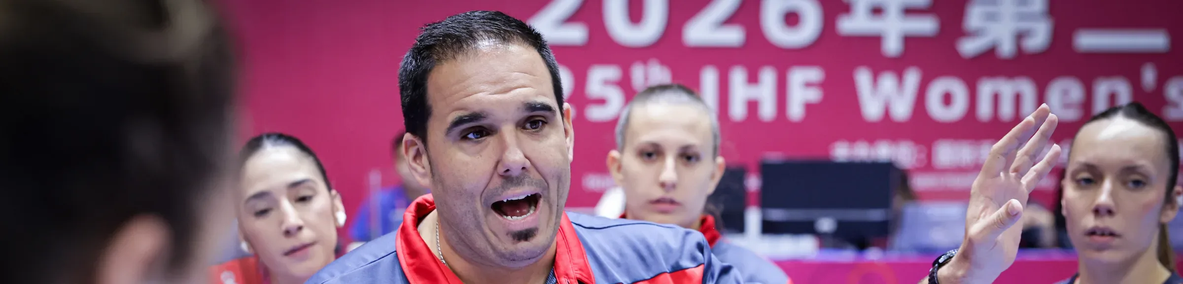 A coach in a red and blue jersey gestures emphatically while holding a play diagram during a women's handball match.