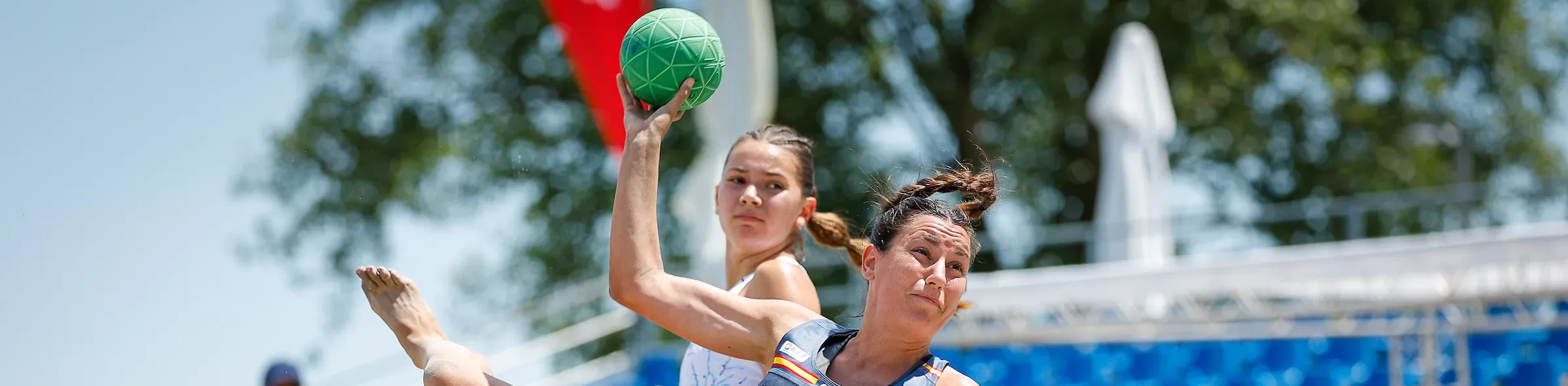 An athlete in a blue uniform leaps mid-air on a sandy court, holding a green ball above their head while another player watches closely.